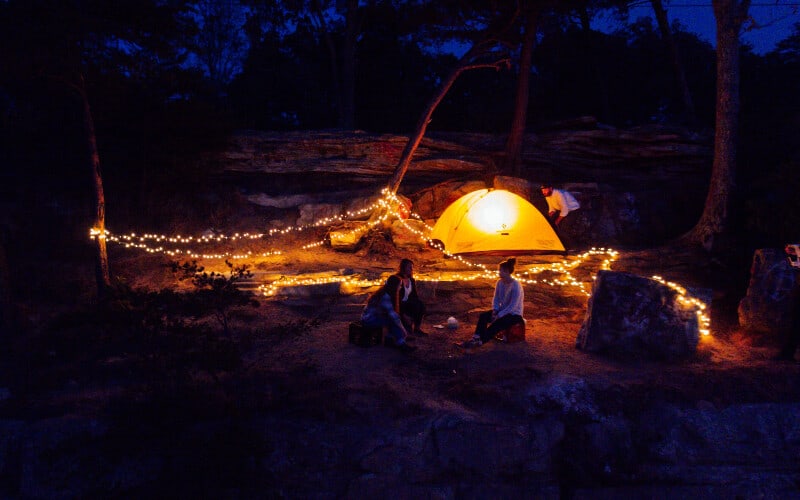 Three people in a campsite surrounded by great campsite lighting ideas: string lights and an illuminated tent.